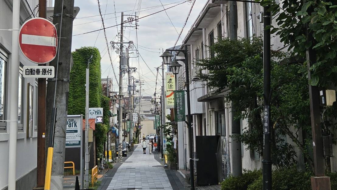 An ordinary shopping street in central Matsumoto at dusk, lined with street lamps, overhead cables and small shopfronts